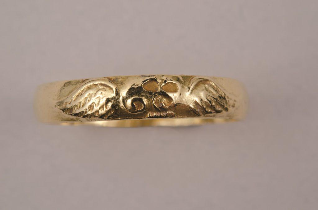 A color photograph of a gold ring with a skull with wings on either side pressed into it. 